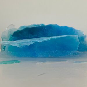 A translucent blue resin sculpture resembling an ice formation, displayed on a white surface with droplets around it.